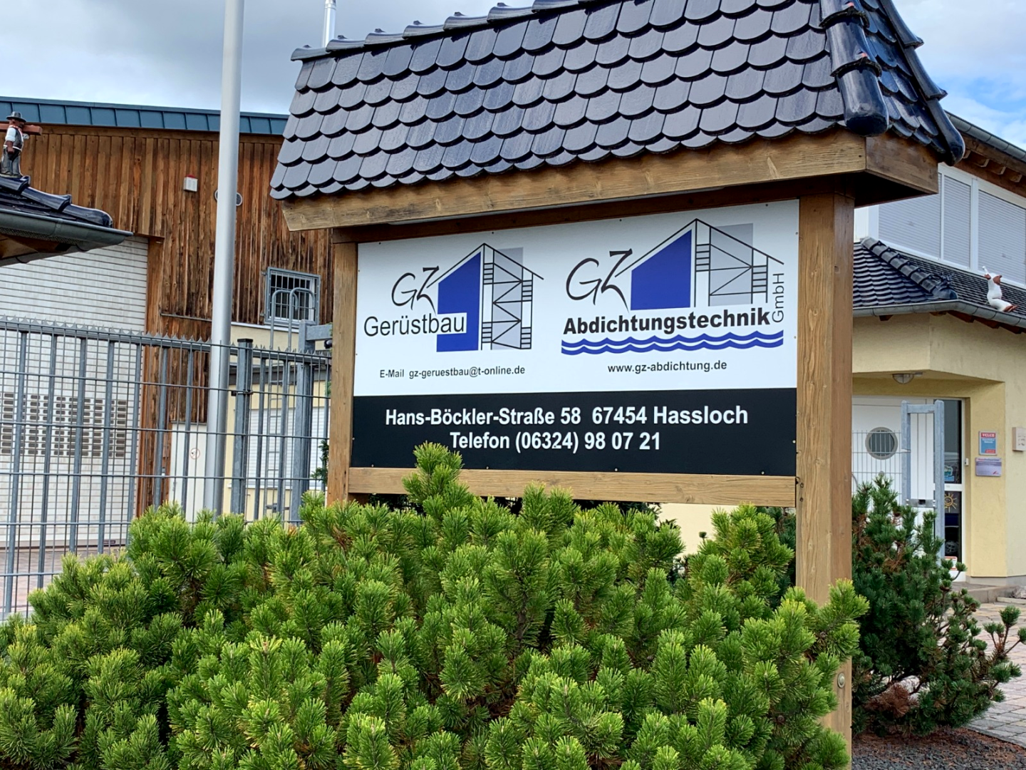 Großes Firmenschild mit Ziegeldach vor Sträuchern an einer Straße