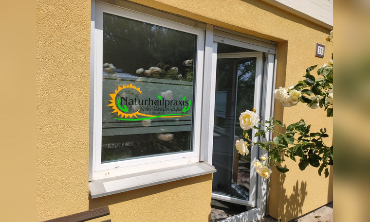 Eingang einer Naturheilpraxis an einer gelben Hauswand mit Fenster, Glastür und weißen Rosen.