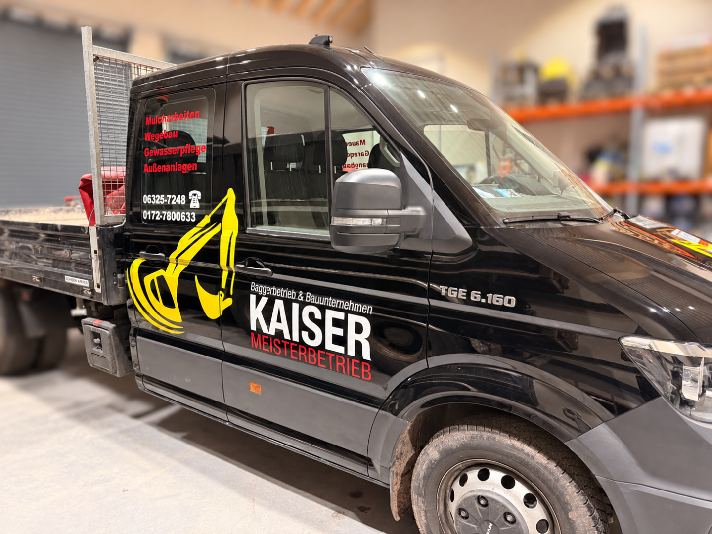 Schwarzer Firmenwagen mit Pritsche in einer Halle, beschriftet mit Kaiser Meisterbetrieb und Baggergrafik.