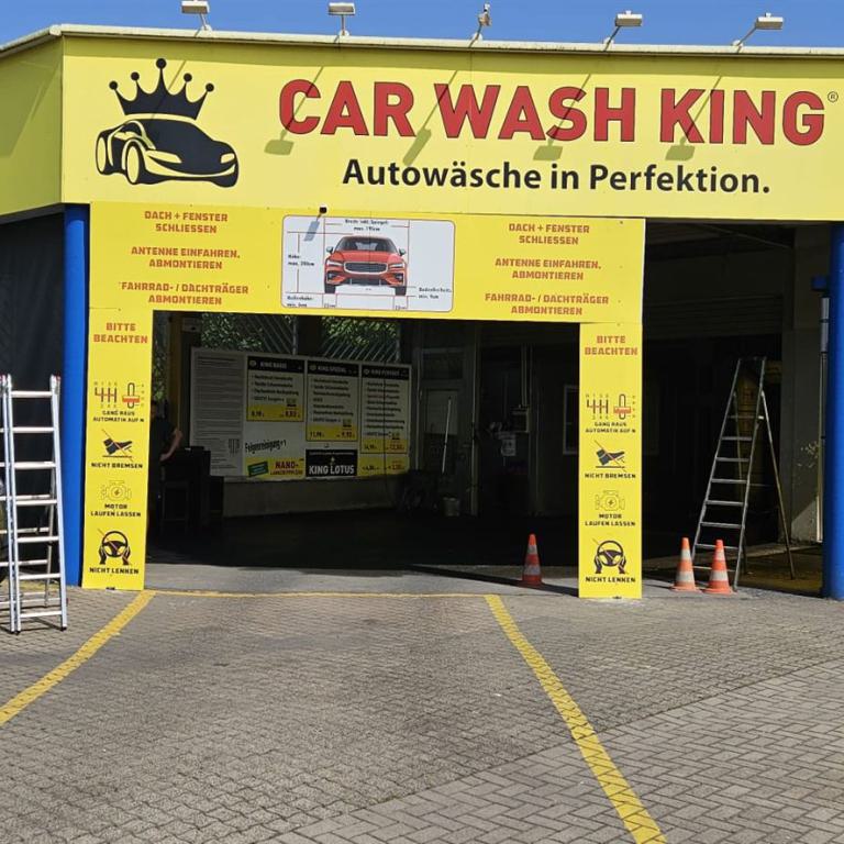 Einfahrt einer gelben Autowaschanlage mit parkenden Autos und Leitern davor.
