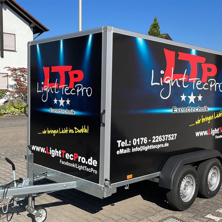 Schwarzer Doppelachsanhänger mit Werbebeschriftung von LightTecPro auf einem Hof vor Wohnhäusern