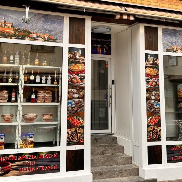 Schaufenster und Eingang eines Balkan-Markts mit Flaschen, Keramik und Werbebildern von Speisen.