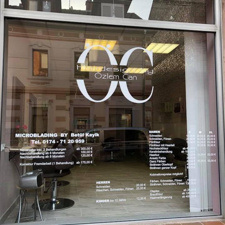 Schaufenster eines Friseursalons mit großem OC-Logo, Preisliste und Blick ins Innere.
