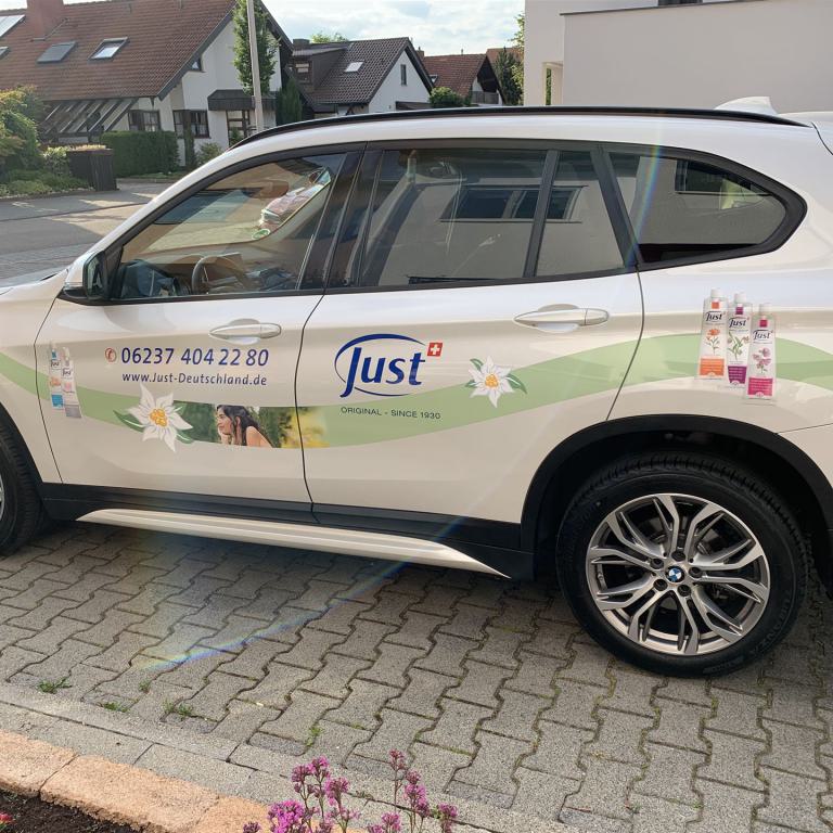 Weißes SUV mit Werbebeklebung der Marke Just auf einem Parkplatz vor Wohnhäusern.